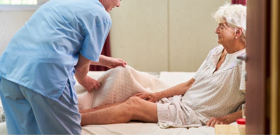 Femme âgée dans son lit, aidée à se lever par une aide soignante. Exemple de soin préventif d'une escarre 